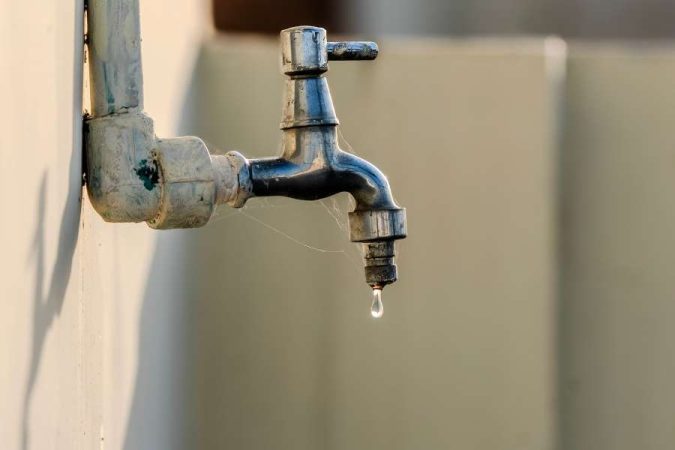 Un vieux robinet en métal fixé sur une canalisation murale laisse échapper une goutte d'eau, nécessitant de réparer un robinet.