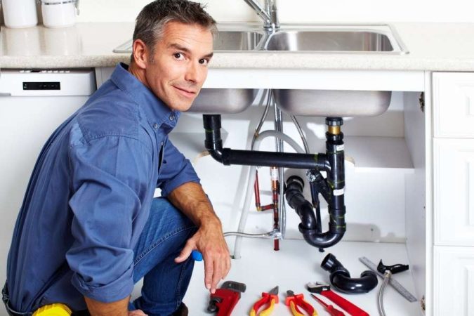 Un homme en chemise bleue s'accroupit sous un évier de cuisine entouré d'outils de plomberie.