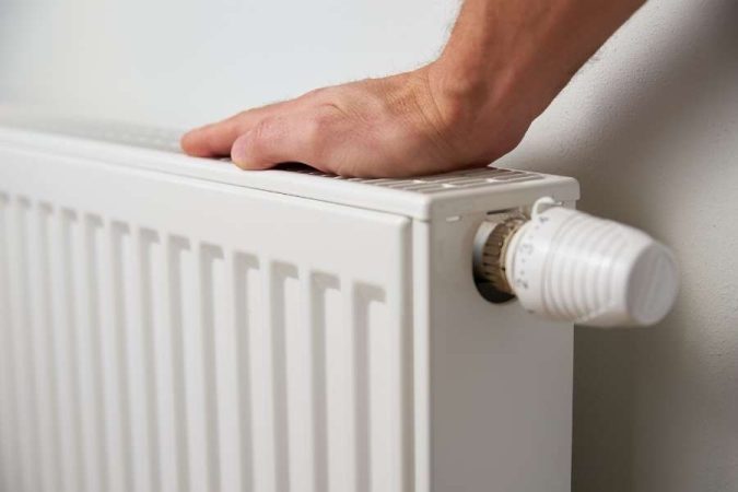 Une main teste la température d'un radiateur blanc qui peut compléter une installation de plinthe chauffante.